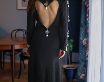 May include: A black long-sleeve dress with a heart-shaped open back. The dress features pearl and rhinestone embellishments on the shoulders and back. The skirt has a pleated design, and the dress is worn with black heels.