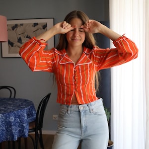 Può includere: Camicia arancione a righe verticali bianche e verdi, colletto bianco e bottoni. Indossata con jeans azzurri. L'outfit ha un'estetica vintage.