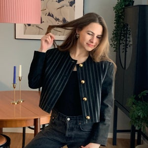 May include: A woman wearing a black and white striped cropped jacket with gold buttons, a black top, and dark wash jeans.