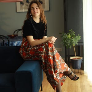 May include: A woman wearing a black velvet top and a long, flowing orange and brown patterned skirt sits on a blue velvet sofa. She is wearing black heels with gold accents.
