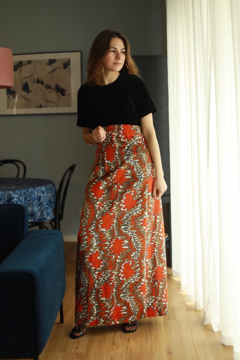 May include: A woman wearing a black short-sleeved shirt and a long orange and brown patterned skirt. The skirt has a swirling, abstract design.