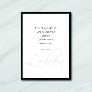 May include: A black framed print with a white background and a quote in German that reads "Es geht nicht darum, was dir im Leben passiert, sondern wie du damit umgehst." The quote is attributed to Monika Creutzburg. The word "Leben" is written in cursive in a light gray color at the bottom of the print.