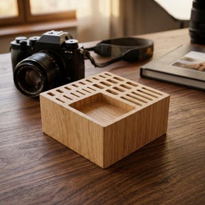 May include: A square light brown wooden organiser block featuring a series of rectangular slots on the top surface and a larger square recessed compartment in the centre, designed for holding small items or tools in a desk workspace.