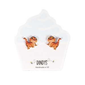 Peut inclure: Une paire de boucles d'oreilles en forme de dragon barbu orange et brun sur une carte blanche en forme de cupcake avec le texte "Dindys Handmade in US".