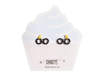 May include: Black and yellow stud earrings shaped like glasses with a lightning bolt detail. The earrings are displayed on a white cupcake-shaped card with the text "Dindys Handmade in US".