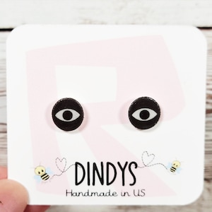 May include: A pair of black and white eye stud earrings on a pink and white card with the text "Dindys Handmade in US".