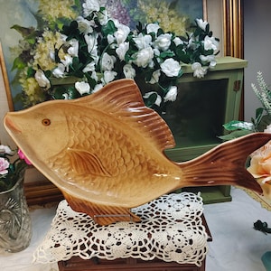 May include: A tan and brown ceramic fish-shaped serving platter with a glossy finish, featuring detailed scale textures, a prominent dorsal fin, and a single dark eye. The platter is displayed in a horizontal orientation with a wide tail fin.