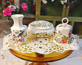 Könnte beinhalten: Ein Set aus weißem Porzellan, bestehend aus einer kleinen Vase, einer runden Dose mit Deckel und einer Glocke, jeweils verziert mit bunten Blumenmustern und Schmetterlingen. Die Stücke stehen auf einer weißen Häkeldecke auf einem bernsteinfarbenen Glasständer.