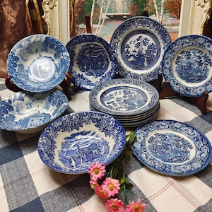 May include: A selection of blue and white ceramic plates and bowls featuring detailed landscape and floral patterns. The tableware is displayed on a checkered tablecloth, with a sprig of pink flowers in the foreground. The plates vary in size and shape.