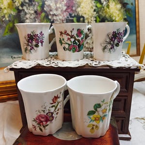 May include: A set of five white porcelain mugs with fluted sides and handles. Each mug features a unique botanical illustration, including purple flowers with ribbons, pink blossoms on branches, and various colorful floral bouquets with roses.