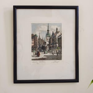May include: Framed print of a European city square with a tall clock tower and detailed architecture. The print is in a black frame with a white mat. The print has text at the bottom.