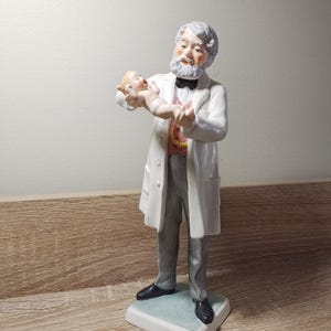 May include: A porcelain figurine of a doctor holding a baby. The doctor is wearing a white coat and a black bow tie. The baby is wearing a white and pink outfit. The figurine is standing on a white base.