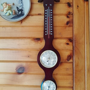 May include: A dark wood-tone weather station with a thermometer, barometer, and hygrometer. The thermometer displays both Fahrenheit and Celsius scales. A decorative plate with a figure playing a horn is to the left.