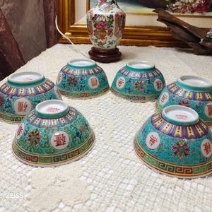 May include: Six small, decorative ceramic bowls with a turquoise background and intricate floral and geometric patterns. Each bowl features a red Chinese character within a white circle. The bowls are arranged on a white, textured surface.