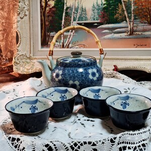 May include: A blue and white teapot with a bamboo handle and four matching teacups. The teapot and cups feature a floral pattern. The teapot is sitting on a white lace tablecloth.