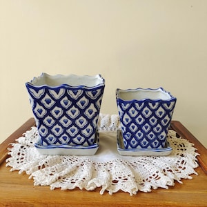May include: Two square ceramic planters with scalloped rims and matching saucers. The planters feature a white base with a repeating blue scale or diamond pattern. The pots are arranged side by side, showcasing a consistent design and finish.