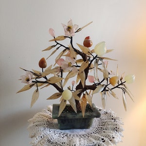 May include: A jade bonsai tree with pink and white flowers and multi-colored gemstone leaves and fruit. The tree is in a green jade pot and is sitting on a white crocheted doily.