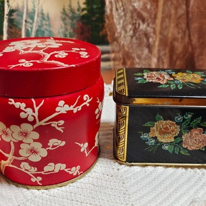 Puede incluir: Dos cajas de hojalata vintage con diseños florales. Una caja es roja con ramas florales blancas y la otra es negra con flores bordadas de colores.