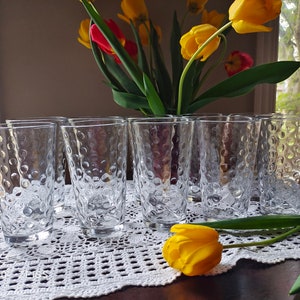 Puede incluir: Juego de ocho vasos transparentes de vidrio con un diseño de burbujas texturizado. Los vasos están dispuestos en una fila sobre un mantel de ganchillo blanco.