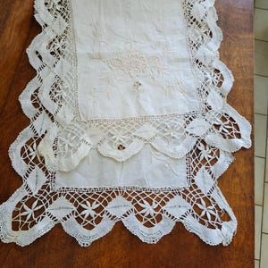 May include: White embroidered table runner with a delicate lace border. The runner features a floral design in the centre.