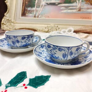 Puede incluir: Dos tazas y platillos de porcelana a juego. Las tazas y los platillos son blancos con un patrón floral y geométrico azul. Las tazas tienen asas y se asientan sobre platillos redondos. El juego está sobre un mantel blanco.