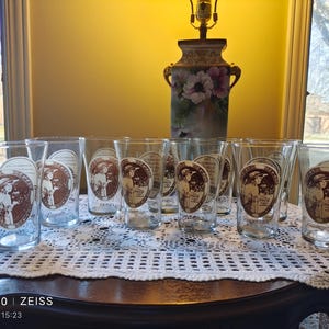 Könnte beinhalten: Satz von zehn klaren Glasbechern mit einem braunen und weißen Design, das eine Frau in einem Vintage-Kleid zeigt. Die Gläser sind auf einer weißen Häkeltischdecke angeordnet.