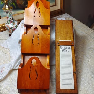 May include: A wooden mail organiser with three tiered pockets and decorative cutouts. Next to it is a wooden grocery list holder with a notepad that reads "Grocery List". Both are made of stained wood.