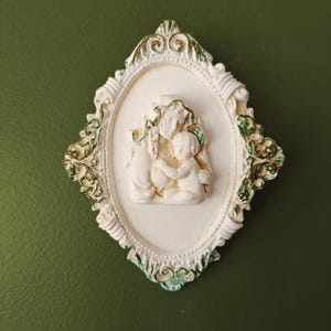 May include: A white oval frame with ornate gold and green details, featuring a white plaster relief sculpture of a mother and child.