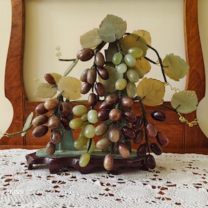 May include: A decorative sculpture of grapes, leaves, and vines. The grapes are in shades of brown, green, and purple. The leaves are a pale green and yellow. The sculpture is mounted on a dark wooden base and set against a wooden frame.