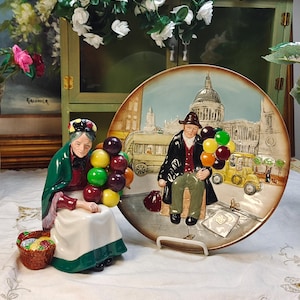 May include: A ceramic figurine of a person wearing a green shawl and white apron holding a cluster of colorful balloons next to a small basket, accompanied by a round decorative plate featuring a relief scene of a balloon seller in London.