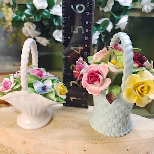 May include: Two porcelain flower baskets with sculpted roses. The left basket is white with a flared base. The right basket is light blue with a woven texture. A black ruler shows the right basket is approximately 9 cm tall.