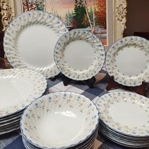 May include: A set of white ceramic dinnerware featuring a floral design. The plates and bowls have a wavy edge with a blue border and are adorned with tiny pink and blue flowers. The collection includes various plate and bowl sizes.
