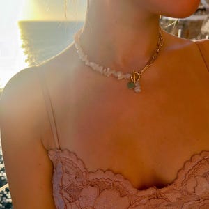 Puede incluir: Un collar con una cadena de cuentas de piedra blanca y una sección de cadena dorada. El collar tiene un cierre dorado y un pequeño dije de corazón verde. La modelo lleva un top de encaje rosa.