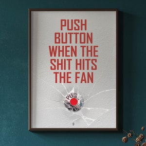 Puede incluir: Una impresión enmarcada con el texto "PUSH BUTTON WHEN THE SHIT HITS THE FAN" en letras rojas. Un botón rojo es visible a través de un panel de vidrio roto.
