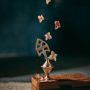 Puede incluir: Un soporte decorativo de metal dorado con una parte superior redonda y tres patas. El soporte está coronado con una pieza de metal decorativa. El soporte está rodeado de delicados pétalos de flores de color rosa claro.