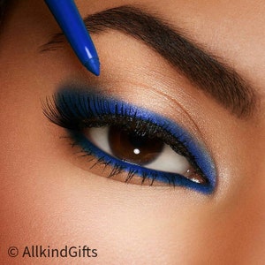 May include: Close-up of an eye with vibrant blue eyeshadow and eyeliner. A blue eyeliner pencil is positioned above the eye. The eye has long, dark eyelashes and a brown iris. The skin around the eye is smooth and highlighted.