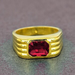 May include: A gold ring featuring a rectangular, deep red gemstone. The ring's band has a ridged design. The gemstone is set within a square bezel, offering a classic and elegant appearance.