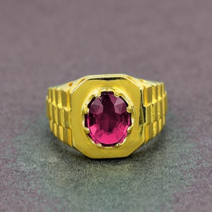 May include: A gold-colored ring with a large, oval, magenta-colored gemstone. The ring has a rectangular design on the band and a geometric setting around the gemstone. The ring is displayed on a dark gray surface.