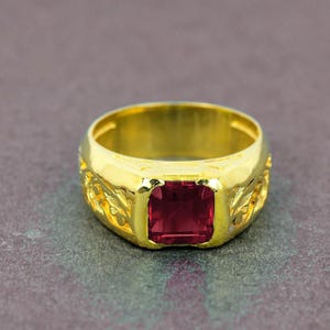 Puede incluir: Un anillo de oro con una gran gema rectangular de color rojo intenso en el centro. La banda del anillo también es dorada y presenta un diseño ornamental en forma de hoja. El anillo está sobre un fondo gris apagado.