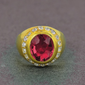May include: A gold ring featuring a large, oval-cut, deep red gemstone at the center. The gemstone is surrounded by a halo of smaller, clear stones. The ring has a thick, polished gold band.
