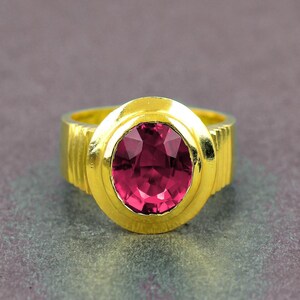 May include: A gold ring with a large, oval-cut, deep pink gemstone. The ring has a thick, textured band and a gold setting that surrounds the gemstone. The ring is set against a muted grey background.