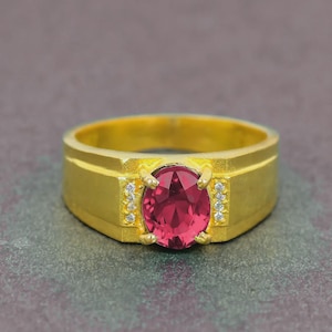 May include: A gold ring featuring a large, oval red gemstone. Small, clear stones are set either side of the central stone. The ring has a rectangular band. The ring is likely made of gold.