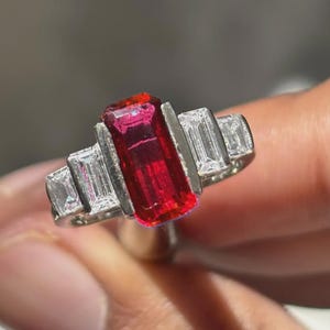 Puede incluir: Un anillo plateado con un diseño llamativo. Presenta una gran gema roja rectangular en el centro, flanqueada por gemas claras rectangulares más pequeñas. El diseño del anillo es moderno, con un contraste de colores audaz.