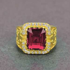 Puede incluir: Un anillo dorado con una gran gema rectangular de color rojo intenso en el centro. La gema está rodeada por un halo de piedras más pequeñas y claras. La banda tiene un diseño grabado y ornamentado.