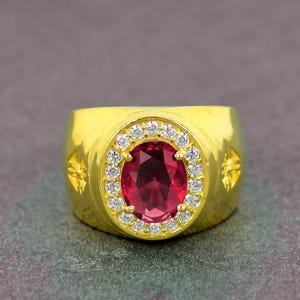 Puede incluir: Un anillo dorado con una gran gema de rubí ovalada, rodeada por un halo de pequeñas gemas claras. El anillo tiene una banda ancha con diseños de estrellas en los lados. Fondo gris oscuro.