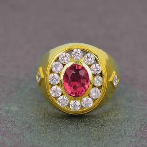 May include: A gold ring featuring a large oval ruby center stone surrounded by a halo of smaller, round, clear gemstones. The ring has a thick band and additional accent stones on the sides. The ring is set against a gray background.