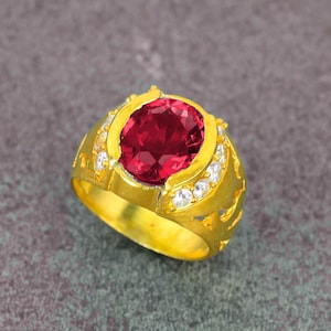 May include: A gold ring featuring a large, oval-cut, deep red gemstone as the centerpiece. The ring is adorned with smaller, clear gemstones and intricate gold filigree detailing on the band. The ring is set against a gray background.