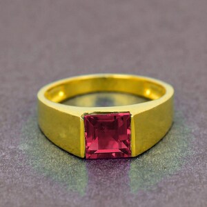 Puede incluir: Un anillo dorado con una gema roja rubí cuadrada. El anillo tiene un diseño sencillo y moderno con una banda gruesa y un engaste de bisel para la gema. El anillo es probablemente de oro y es una pieza de joyería.