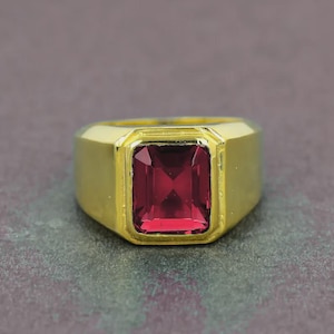 May include: A gold ring featuring a rectangular, deep red gemstone. The ring has a thick band and a classic design. The gemstone is set in a square bezel. The ring is likely made of gold.