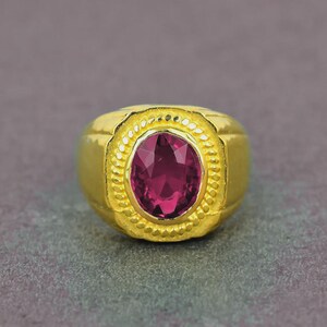 May include: A gold ring featuring an oval, deep red gemstone. The ring has a textured, beaded border around the gemstone. The band is solid, polished gold. The ring is set against a grey background.